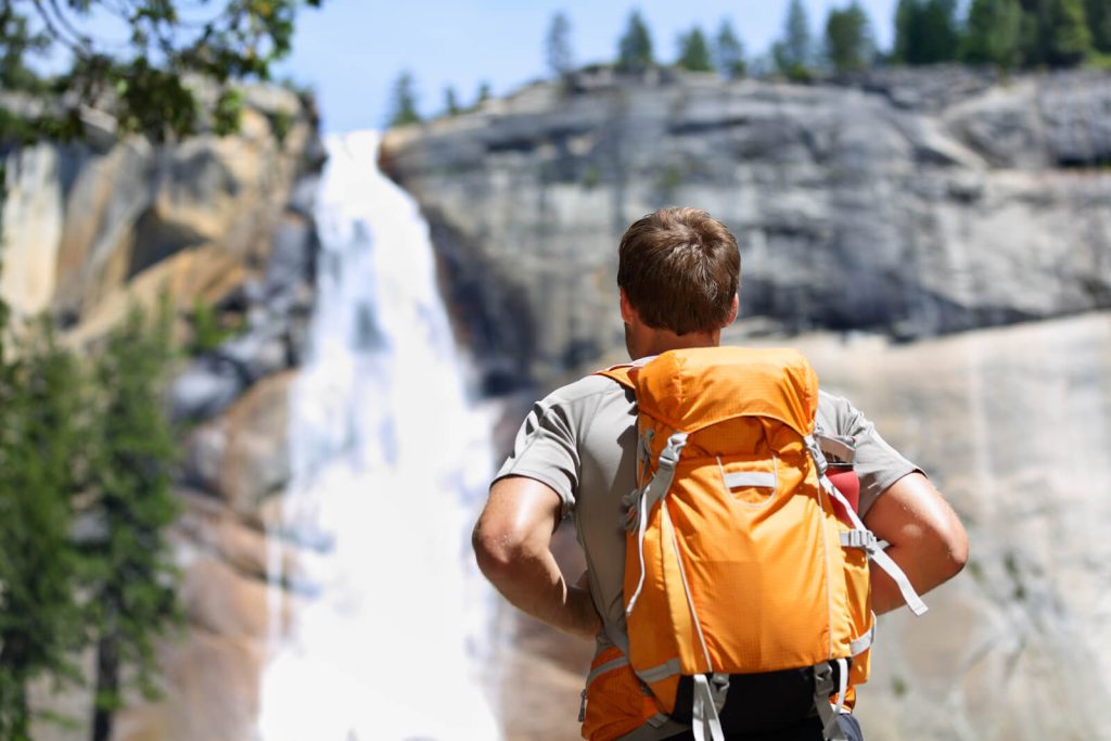 Man with back to the camera wearing orange backpack looking at waterfall in Yosemite in summer