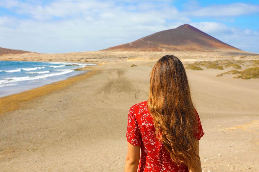 Woman in red dress wiht back to camera walking on beach with volcano in distance