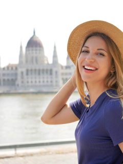 Woman in blue t shirt and hat with the Budapest Parliament building in background across the river