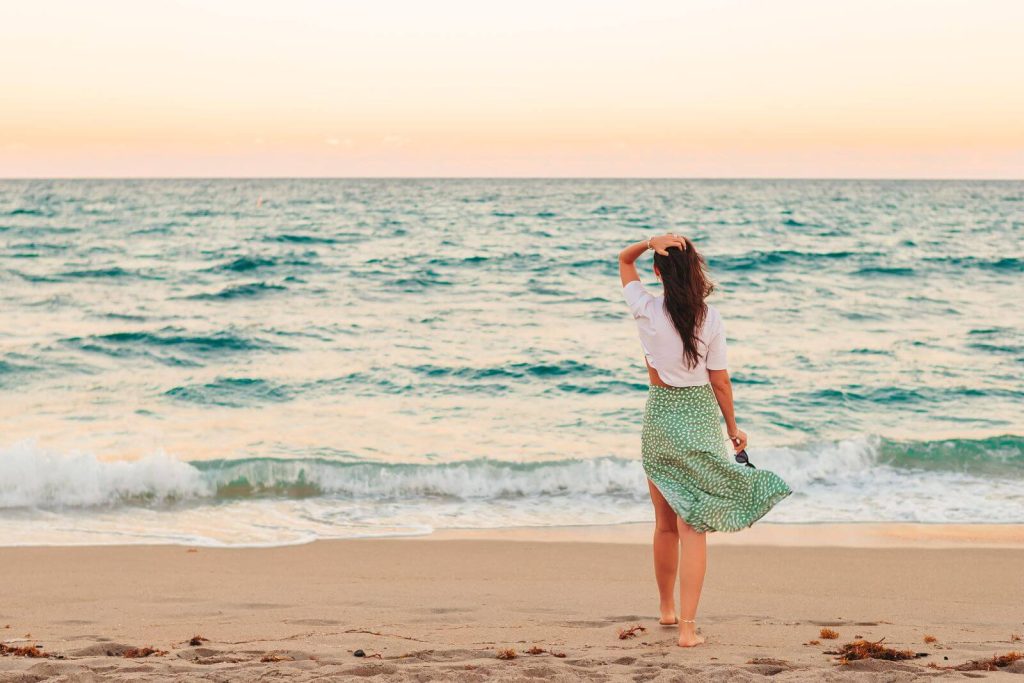 Woman with her back to camera looking at sea on beach in a white top and green skirt