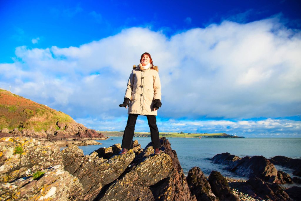Woman in Winter clothes stood on rock with sea in background in Ireland