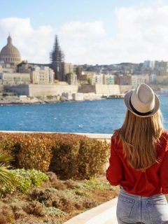 Woman with her back to the camera in red top and straw hat with skyline of Valletta in Malta in background across walter